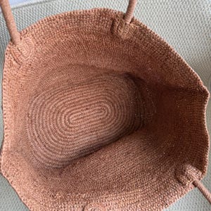 Pode incluir: Uma sacola tote tecida em um tom marrom quente. A sacola apresenta um interior espa&ccedil;oso com uma base oval e al&ccedil;as resistentes. A textura da sacola &eacute; vis&iacute;vel, mostrando as fibras naturais usadas em sua constru&ccedil;&atilde;o. A sacola &eacute; projetada para transportar itens.