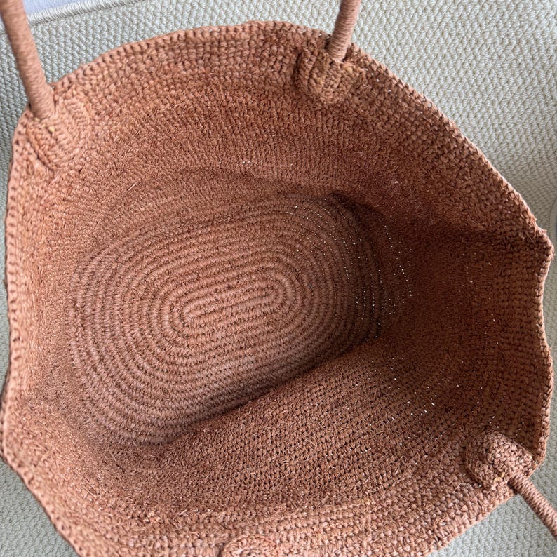 Pode incluir: Uma sacola tote tecida em um tom marrom quente. A sacola apresenta um interior espa&ccedil;oso com uma base oval e al&ccedil;as resistentes. A textura da sacola &eacute; vis&iacute;vel, mostrando as fibras naturais usadas em sua constru&ccedil;&atilde;o. A sacola &eacute; projetada para transportar itens.