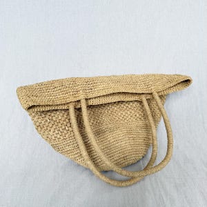 May include: A beige woven straw tote bag with two handles and a folded-over top. The bag's texture reveals the intricate weave of the straw material. The bag is empty and rests on a light grey surface. The bag is a natural colour.
