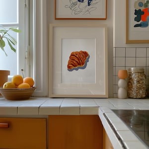 Puede incluir: Impresión enmarcada de un cruasán sobre fondo blanco. La obra de arte está en un marco de madera clara y se exhibe en una encimera de cocina. También se ven un cuenco de naranjas y un objeto decorativo.
