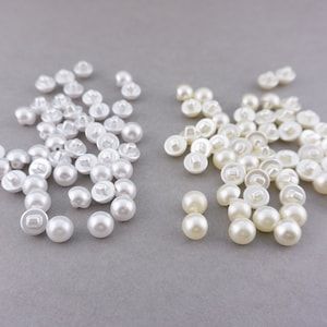 Op de afbeelding: Een grote stapel witte parelknopen en een kleinere stapel crèmekleurige parelknopen.