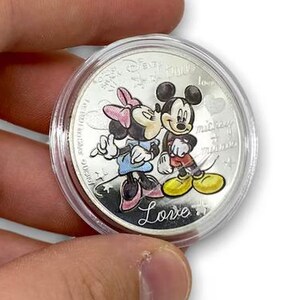 Könnte beinhalten: Eine Silbermünze mit Mickey und Minnie Mouse in einer liebevollen Umarmung, eingebettet in einer transparenten Schutzhülle. Auf der Münze steht das Wort "Love".