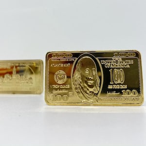 May include: Two gold-coloured bars. The larger bar is shaped like a $100 bill, with the text "100" and "United States of America." The smaller bar has an image of a building. Both bars are on a white surface.