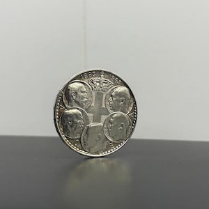 Può includere: Moneta d'argento con più profili di persone, con gli anni 1863 e 1963 incisi in alto. La moneta ha un disegno a croce al centro e un bordo dettagliato.