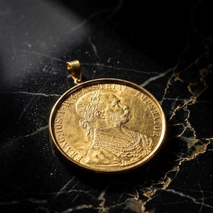 Puede incluir: Colgante dorado con un perfil detallado de un hombre y texto. El colgante tiene un lazo para una cadena. Descansa sobre una superficie oscura y veteada.