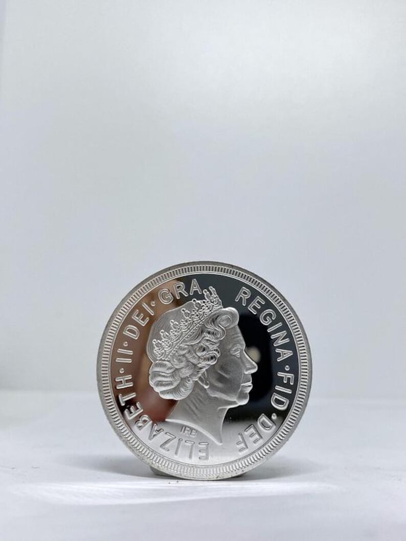 May include: A silver coin featuring a profile of a crowned person, with the inscription "ELIZABETH II DEI GRA REG F.D." around the edge. The coin has a reflective surface and is set against a plain white background.