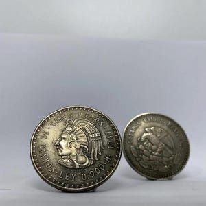 Könnte beinhalten: Zwei silberne mexikanische Münzen. Die größere Münze zeigt das Profil einer Person mit Kopfschmuck und Text am Rand. Die kleinere Münze zeigt einen Adler mit Text. Beide Münzen sind vor einem weißen Hintergrund platziert.