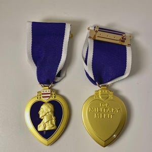 Pode incluir: Duas medalhas Purple Heart douradas com fitas listradas azuis e brancas. Uma apresenta um perfil de George Washington, a outra diz "FOR MILITARY MERIT". As medalhas têm formato de coração.