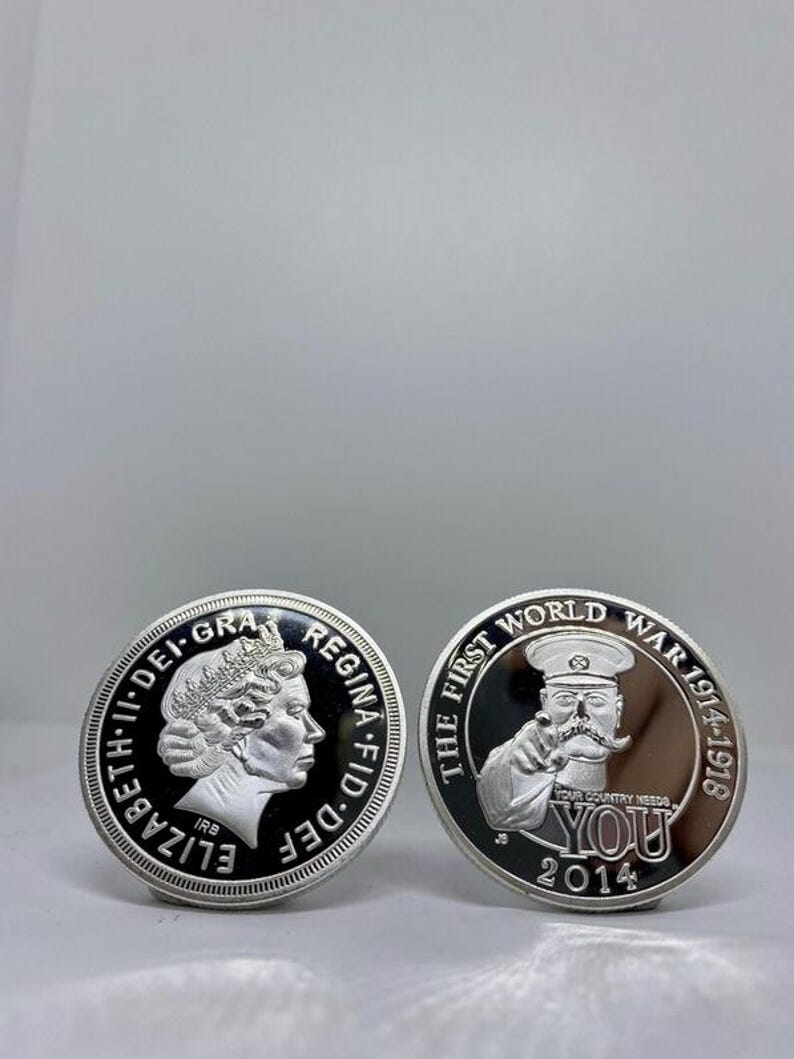 May include: Two silver coins. One features a profile of Queen Elizabeth II and the text "ELIZABETH II DEI GRA REGINA FID DEF". The other coin has a man pointing with the text "THE FIRST WORLD WAR 1914-1918 YOUR COUNTRY NEEDS YOU 2014".