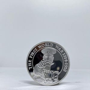 May include: A silver coin featuring a man in a military cap pointing his finger. The coin reads "THE FIRST WORLD WAR 1914-1918" around the edge, with "YOUR COUNTRY NEEDS YOU" and the year 2014 below the image.