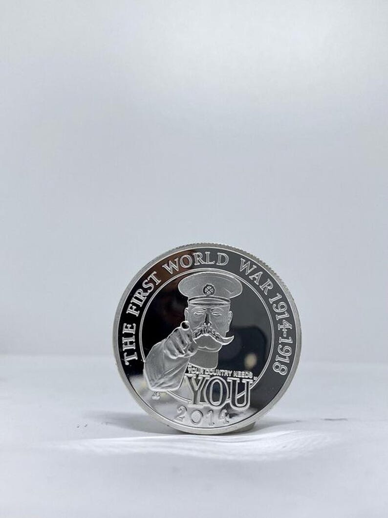 May include: A silver coin featuring a man in a military cap pointing his finger. The coin reads "THE FIRST WORLD WAR 1914-1918" around the edge, with "YOUR COUNTRY NEEDS YOU" and the year 2014 below the image.