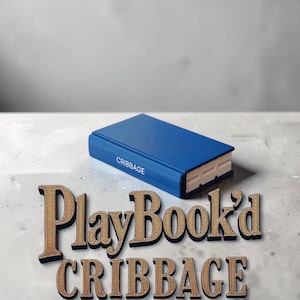 May include: A rectangular blue game board shaped like a book with the word CRIBGAGE printed on the spine. The white interior of the board is visible at the end. Below the object, the text PlayBook'd CRIBGAGE is displayed in large gold letters.