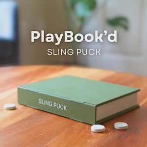 Pode incluir: Um jogo Sling Puck verde sobre uma mesa de madeira. A caixa do jogo é retangular e está rotulada como "SLING PUCK". Três discos brancos estão espalhados ao redor da caixa. O texto "PlayBook'd" está na parte superior da imagem.