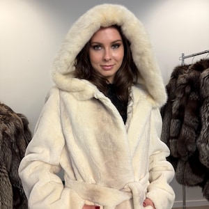 May include: A cream-coloured faux fur coat with a hood. The coat features a wrap-around design and a matching belt. The hood frames the face, and the coat looks soft and warm. Other fur coats are visible in the background.
