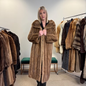 Könnte beinhalten: Ein langer, brauner Pelzmantel mit breitem Kragen. Der Mantel reicht bis zu den Knien und scheint aus einem weichen, luxuriösen Material zu bestehen. Weitere Pelzmäntel hängen auf Kleiderständern im Hintergrund.