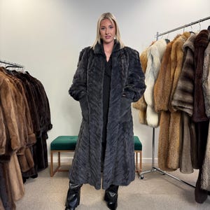 Peut inclure: Un long manteau de fourrure gris foncé avec un motif texturé. Le manteau a un col et des manches longues. L'image montre une femme portant le manteau, avec d'autres manteaux de fourrure en arrière-plan. Des bottes noires complètent le look.