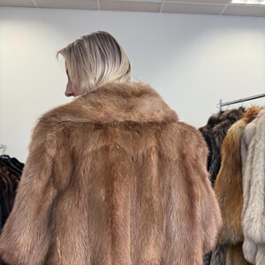 Puede incluir: Un abrigo de piel marr&oacute;n claro con cuello ancho y mangas cortas. El abrigo se muestra sobre una persona, con otros abrigos de piel visibles en el fondo. La piel parece suave y lujosa, con un color natural.