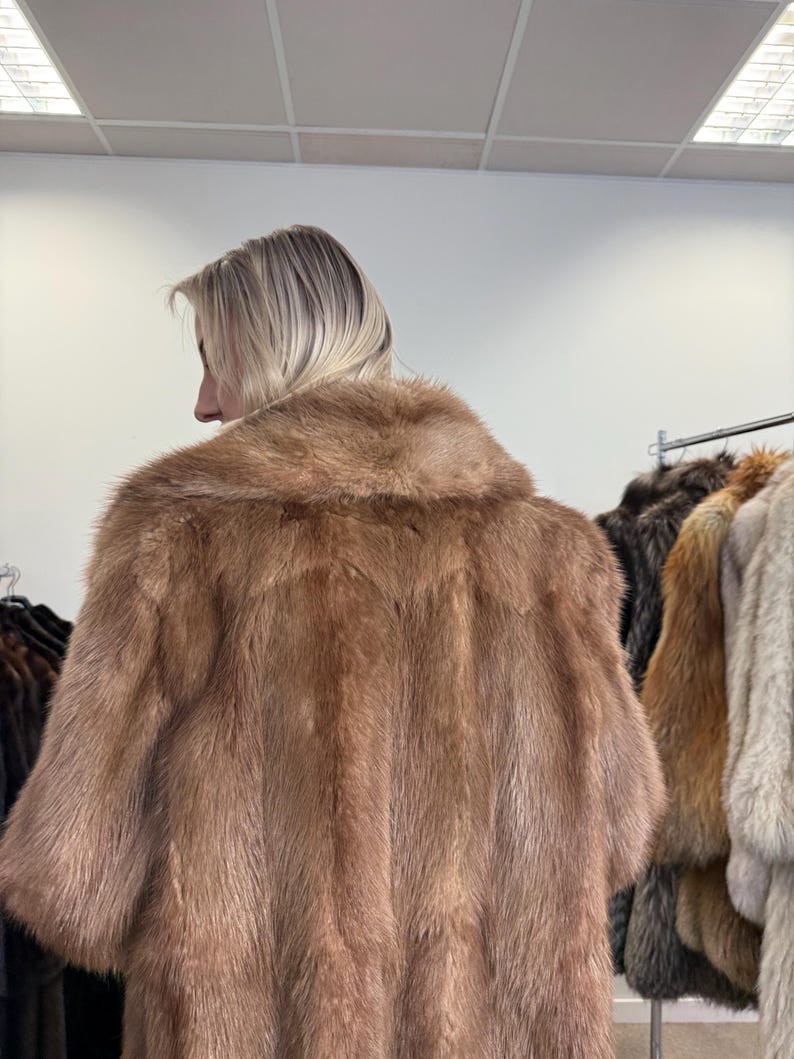Puede incluir: Un abrigo de piel marr&oacute;n claro con cuello ancho y mangas cortas. El abrigo se muestra sobre una persona, con otros abrigos de piel visibles en el fondo. La piel parece suave y lujosa, con un color natural.