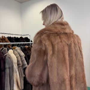 Puede incluir: Una persona lleva un abrigo largo de piel de color marr&oacute;n claro con un gran cuello. El abrigo es el punto focal, con una apariencia suave y texturizada. Otros abrigos de piel de varios colores cuelgan en un estante en el fondo.