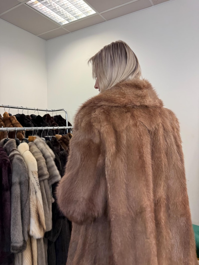 Puede incluir: Una persona lleva un abrigo largo de piel de color marr&oacute;n claro con un gran cuello. El abrigo es el punto focal, con una apariencia suave y texturizada. Otros abrigos de piel de varios colores cuelgan en un estante en el fondo.