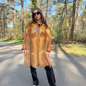 May include: A long brown faux fur coat with a button closure. The coat has a fur collar and is worn over black trousers and white trainers.