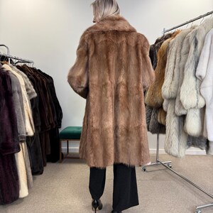 Puede incluir: Un abrigo largo de piel marr&oacute;n con un cuello ancho y mangas tres cuartos. El abrigo llega a la rodilla y lo lleva una persona con pantalones negros y tacones altos. Otros abrigos de piel cuelgan en percheros en el fondo.