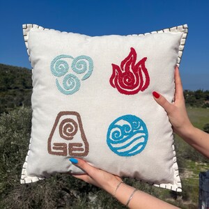 May include: White cushion with embroidered symbols of the four elements: air, fire, earth, and water. The symbols are in blue, red, brown, and light blue.