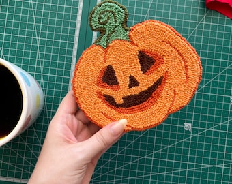 May include: Handmade orange and brown pumpkin-shaped coaster with a smiling jack-o'-lantern face and green stem. The coaster is made of textured yarn and is a festive Halloween decoration.
