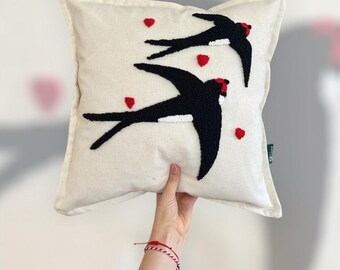 May include: Cream-colored square pillow featuring two black swallows with red accents and small red heart shapes. The swallows have white details and are embroidered on the pillow. The pillow is held by a person.