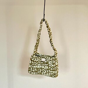 Op de afbeelding: Een klein groen en wit gecrochet handtasje met een korte schouderband die aan een haak hangt.