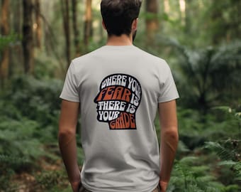 May include: A white t-shirt with a graphic of a head with the text "Where your fear is there is your grade" printed on the back.