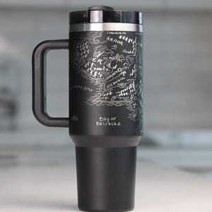 May include: Black travel mug with a silver map design and a black handle. The map includes text such as "Eriador" and "Bay of Belfalas". The mug has a black lid and a silver rim.