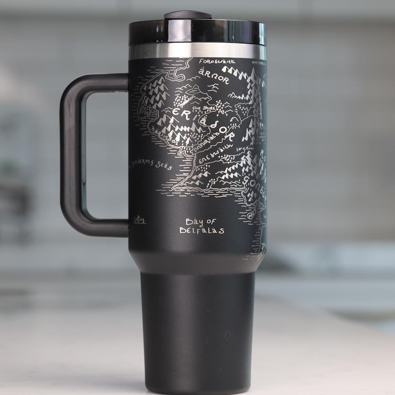 May include: Black travel mug with a silver map design and a black handle. The map includes text such as "Eriador" and "Bay of Belfalas". The mug has a black lid and a silver rim.