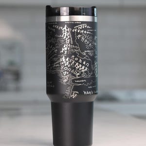 May include: Black travel mug with a silver map design. The map includes text such as "Gondor" and "Rohan". The mug has a black lid and a silver rim.
