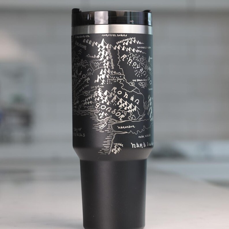 May include: Black travel mug with a silver map design. The map includes text such as "Gondor" and "Rohan". The mug has a black lid and a silver rim.