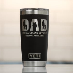 May include: Black insulated tumbler with a silver rim and lid. The tumbler features the word "DAD" with silhouettes of a father and children. Below the word "DAD" is the text "DEDICATED AND DEVOTED TO ELIANA AND KARINA". The Yeti logo is at the bottom.