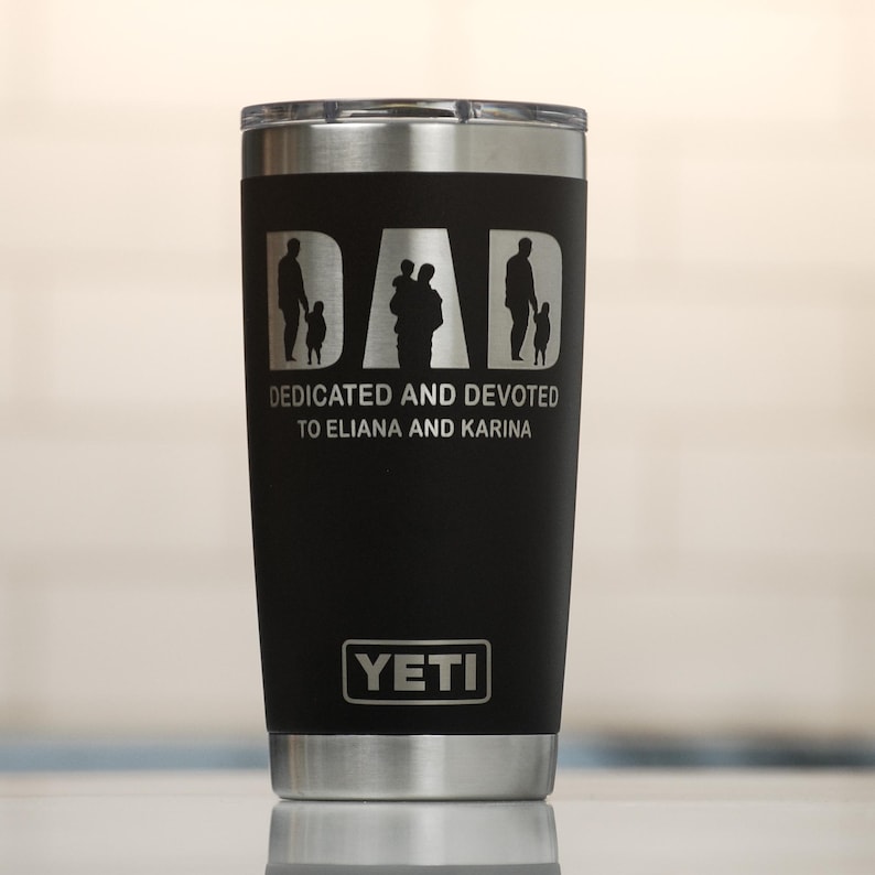 May include: Black insulated tumbler with a silver rim and lid. The tumbler features the word "DAD" with silhouettes of a father and children. Below the word "DAD" is the text "DEDICATED AND DEVOTED TO ELIANA AND KARINA". The Yeti logo is at the bottom.
