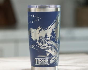 May include: A navy blue stainless steel tumbler with a laser-engraved design featuring a jumping fish, pine trees, mountains, and a flock of birds. The text GONE FISHING is printed in a rectangular frame at the bottom of the cylindrical cup.