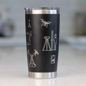 Pode incluir: Um copo de aço inoxidável preto fosco com ícones de linhas brancas gravados a laser relacionados com topografia. O design inclui um drone, um tripé com estação total, uma pessoa com mira topográfica, satélites e uma prancheta.