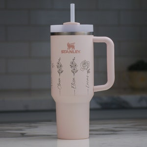 May include: A light pink Stanley tumbler with a handle and straw. The tumbler features the Stanley logo and floral illustrations with names: Clara, Eliza, and Lucas. The lid is white and the handle is the same pink color as the tumbler.