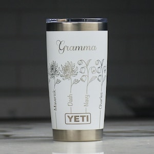 May include: A white Yeti travel mug with a silver lid and base. The word "Gramma" is written in cursive above a floral design. Below the flowers are the names "Maverick", "Dash", "Navy", and "Charles". The Yeti logo is centered below the names.