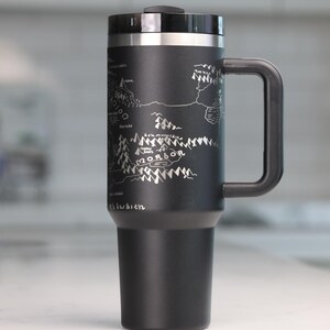 May include: Black travel mug with a silver map design and a black handle. The map features the word "Mordor" and other text. The mug has a black lid and a silver rim. The mug is approximately 8 inches tall.
