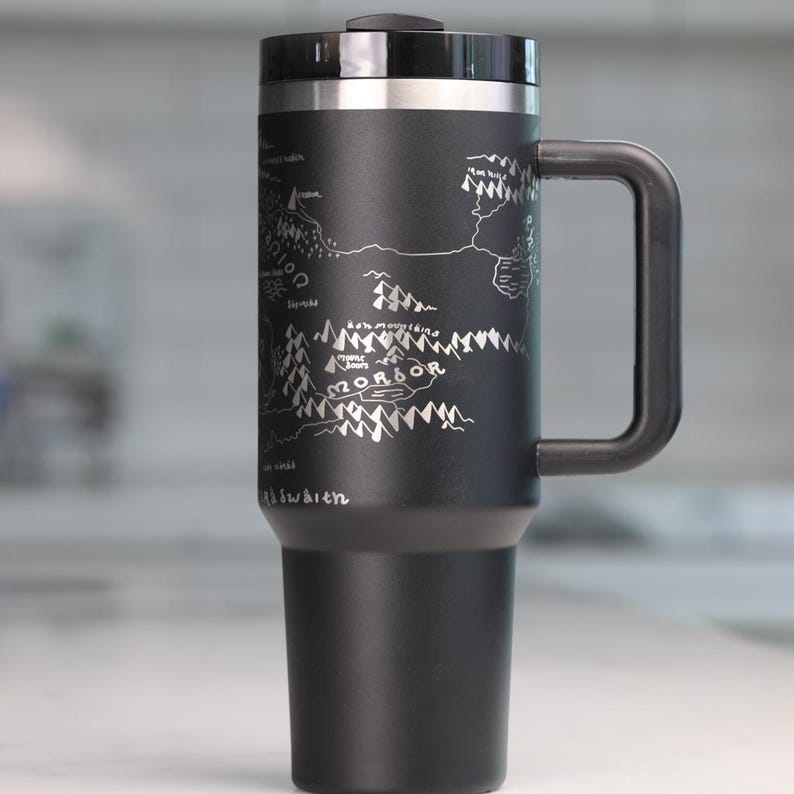 May include: Black travel mug with a silver map design and a black handle. The map features the word "Mordor" and other text. The mug has a black lid and a silver rim. The mug is approximately 8 inches tall.