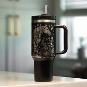 May include: Black insulated travel mug featuring a silver horse and floral pattern. The mug has a black handle, lid, and base, and includes a white straw. The design is etched into the black surface.