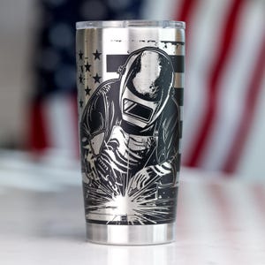 May include: A stainless steel tumbler featuring a black laser-etched graphic of a welder wearing a protective helmet and apron, actively welding with visible sparks. The background design incorporates a stylized American flag with stars and stripes.