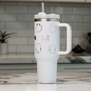 Puede incluir: Taza de viaje de acero inoxidable blanca con asa negra y tapa blanca. La taza tiene un dise&ntilde;o de varios gatos con diferentes expresiones.
