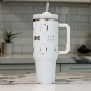 Puede incluir: Taza de viaje de acero inoxidable blanca con asa negra y pajita. La taza tiene un fondo blanco con un patr&oacute;n de gatos blancos y negros.