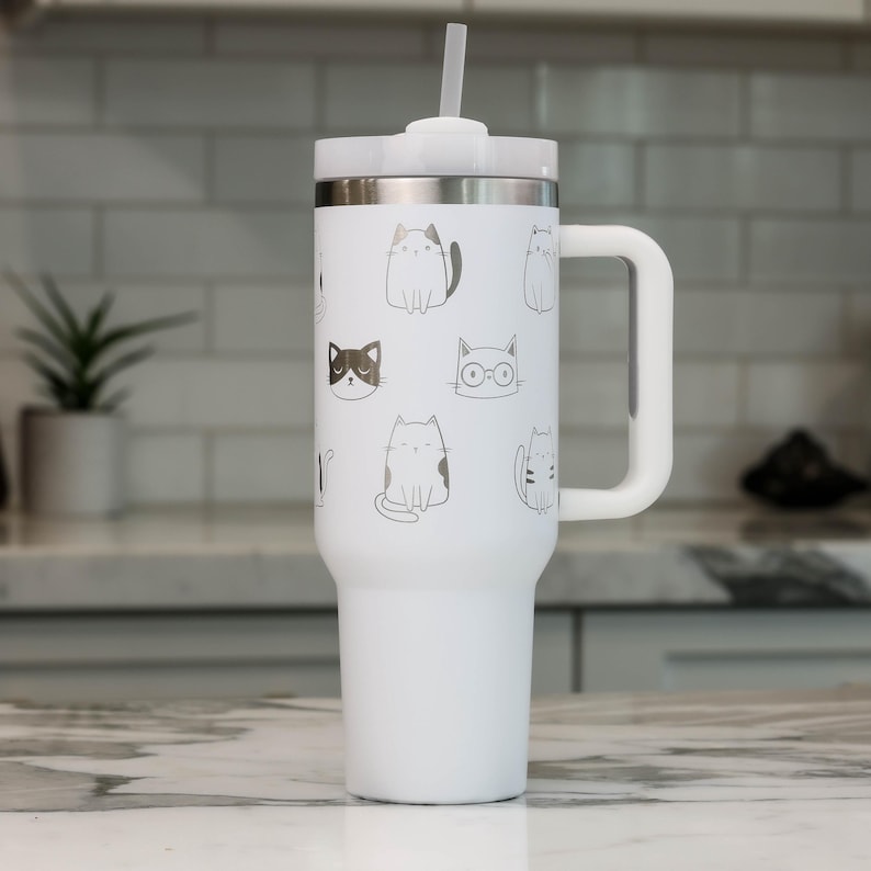 Puede incluir: Taza de viaje de acero inoxidable blanca con asa negra y pajita. La taza tiene un fondo blanco con un patr&oacute;n de gatos blancos y negros.