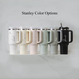 Puede incluir: Una fila de siete termos de viaje Stanley en diferentes colores, incluyendo negro, crema, rosa, verde y gris. Los termos tienen un asa y una tapa con una pajita. El texto "Stanley Color Options" est&aacute; en la parte superior de la imagen.
