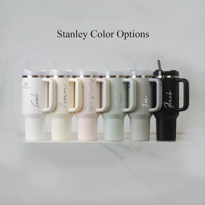 Puede incluir: Una fila de siete termos de viaje Stanley en diferentes colores, incluyendo negro, crema, rosa, verde y gris. Los termos tienen un asa y una tapa con una pajita. El texto "Stanley Color Options" est&aacute; en la parte superior de la imagen.